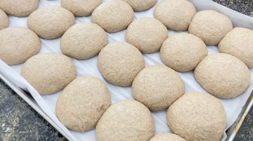 Well proofed sourdough buns resting on a cookie sheet.  They are ready to bake.